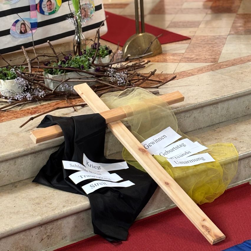 Ein hölzerne Kreuz mit schwarzem Tuch liegt auf einer Kirchenstufe. Daneben sind gelber Stoff und kleine Pflanzen zu sehen. Das Kreuz hat Etiketten wie 'Gewinnen', 'Geburtstag' und 'Umarmung'. Dahinter befindet sich ein roter Teppich und ein weißer Stoff mit Bildern ziert den Hintergrund.