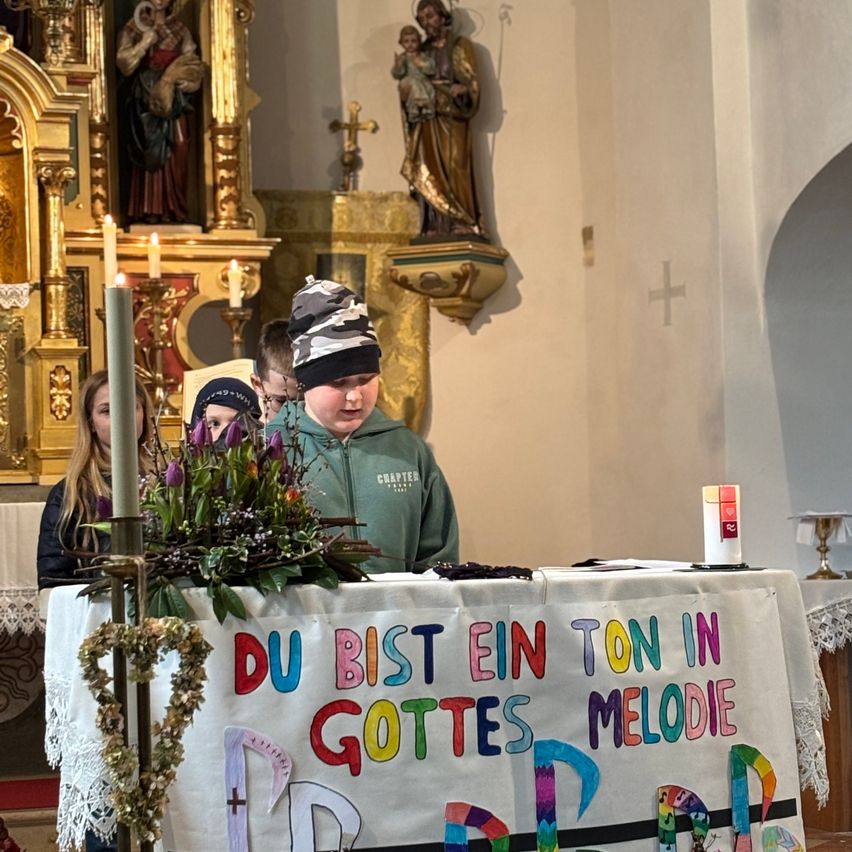 Ein Junge steht an einem Podium mit einem weißen Tuch vor sich. Dahinter stehen zwei andere Kinder vor einem Kerzenhalter. Ein Banner mit buntem Text steht 'Du bist ein Ton in Gottes Melodie'.