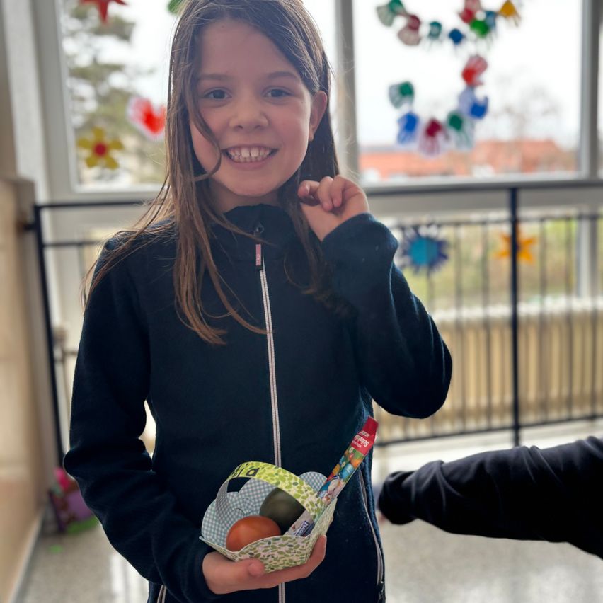 Ein Mädchen in einer schwarzen Jacke hält einen Osterkorb mit Schokoladeneiern und Süßigkeiten. Sie lächelt und steht vor einem Fenster mit einer bunten Handabdruckdekoration.
