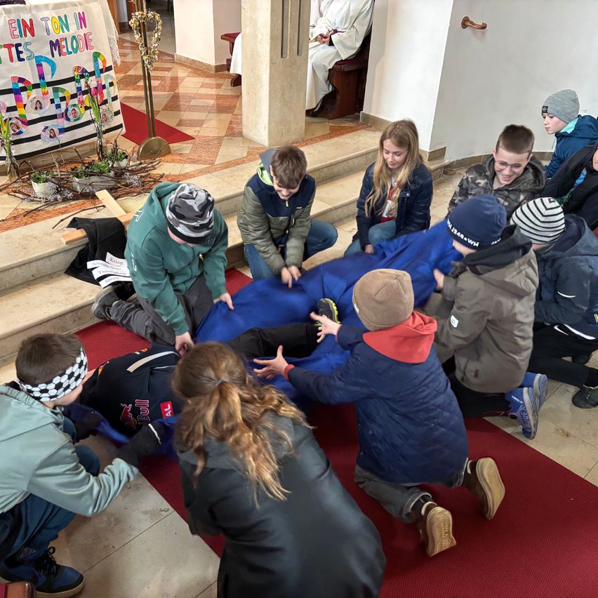 In einer Kirche versammeln sich eine Gruppe von Kindern um einen blauen Stoff. Sie scheinen einen Ritus durchzuführen. Ein Banner mit Text hängt im Hintergrund.