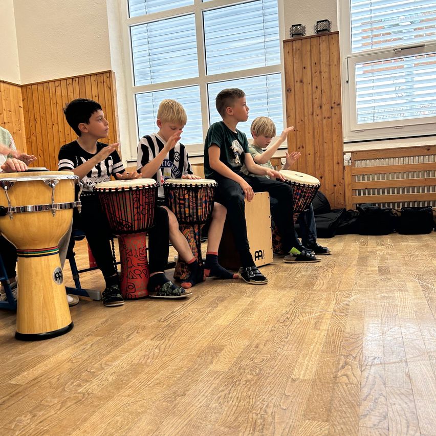Fünf Kinder sitzen auf einem Holzboden, jedes hält eine Trommel und einen Drumstick. Sie scheinen zu üben oder aufzutreten. Holzwände und Fenster sind im Hintergrund.