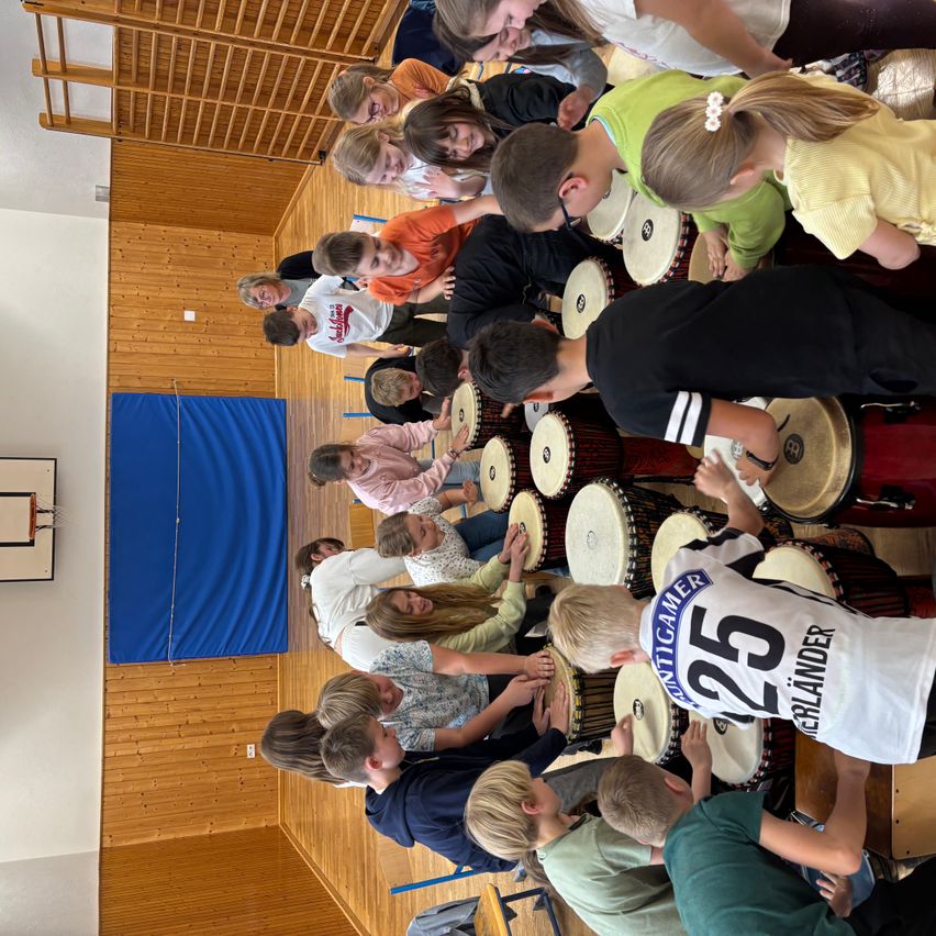 Eine Gruppe von Kindern und Erwachsenen spielt in einem Raum gemeinsam Trommeln. Die Kinder tragen verschiedenfarbige T-Shirts.