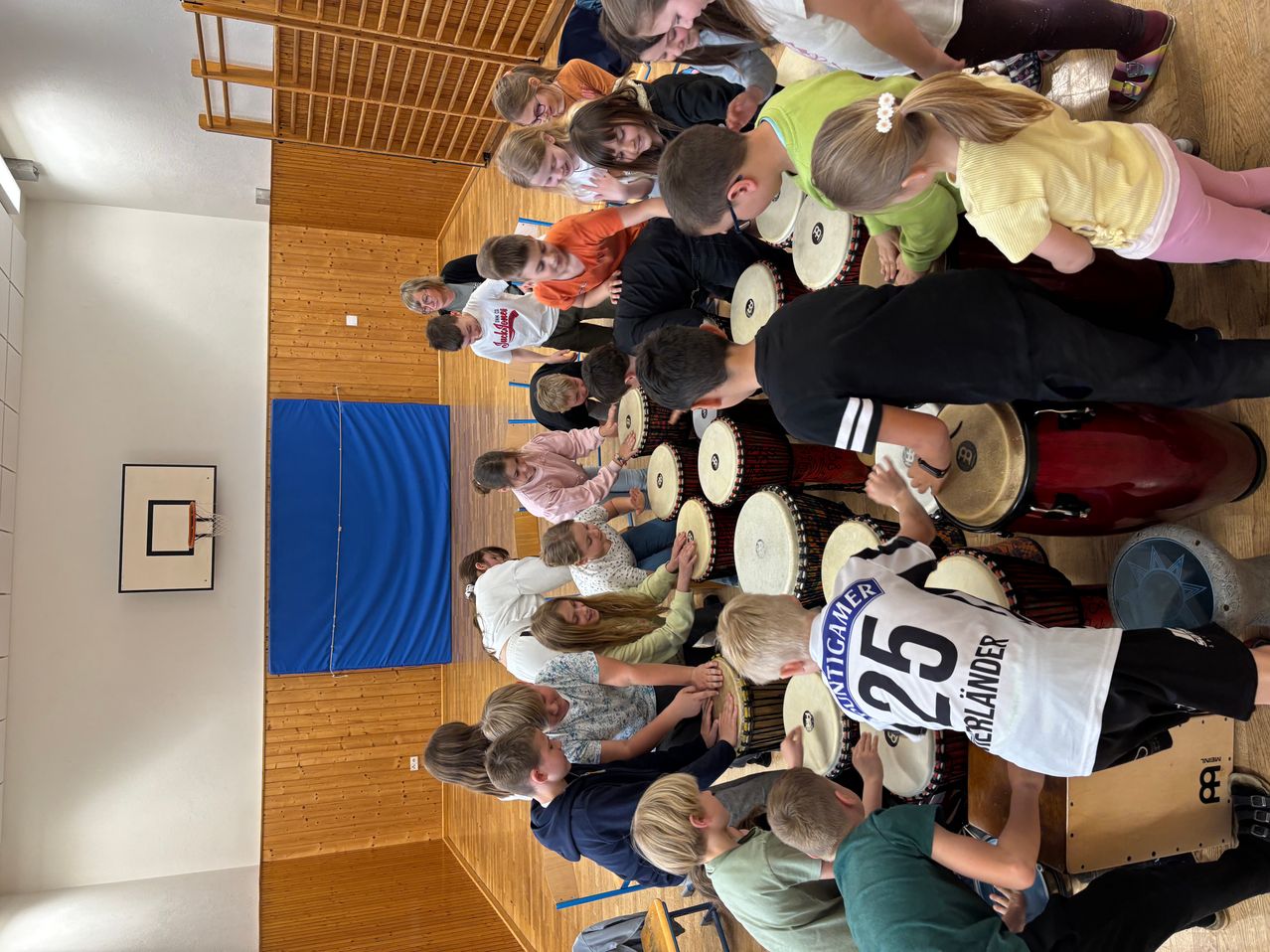 Eine Gruppe von Kindern und Erwachsenen spielt in einem Raum gemeinsam Trommeln. Die Kinder tragen verschiedenfarbige T-Shirts.