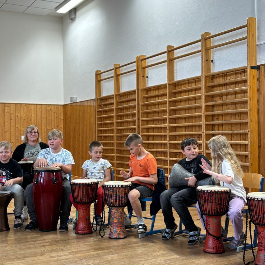 Eine Gruppe von Kindern in einem Raum spielt Trommeln und lächelt. Sie sitzen auf Stühlen in einer Reihe mit Holzwänden dahinter.