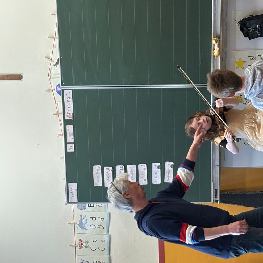 Ein junger Junge und ein Mädchen spielen Violinen. Ein älterer Mann hilft ihnen. Die Wand hat Etiketten und eine grüne Tafel.