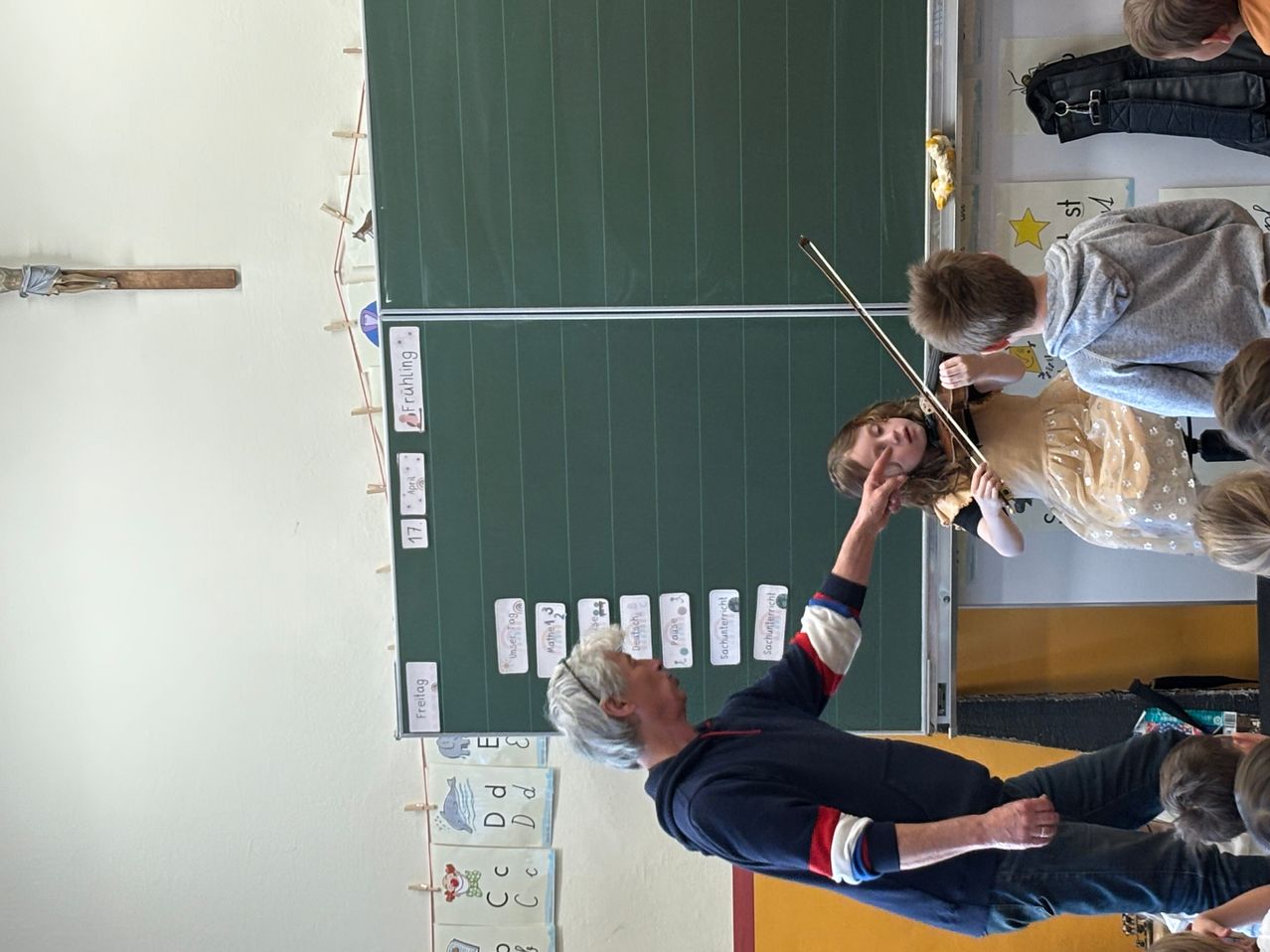 Ein junger Junge und ein Mädchen spielen Violinen. Ein älterer Mann hilft ihnen. Die Wand hat Etiketten und eine grüne Tafel.