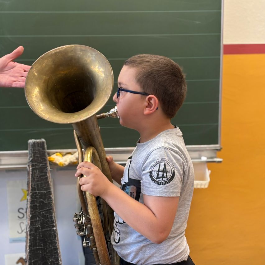 Ein Junge spielt ein Blechblasinstrument im Klassenzimmer, mit einer Tafel im Hintergrund und einer Hand auf der linken Seite.