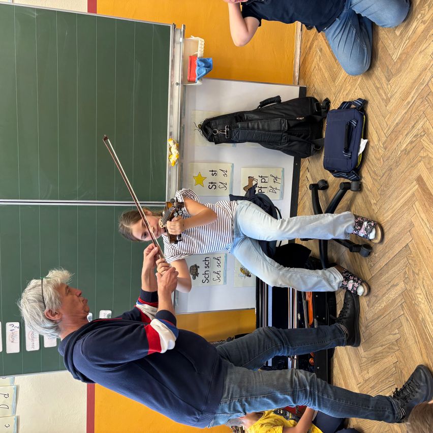 Ein Mann hilft einem jungen Mädchen, Geige zu spielen, in einem Klassenzimmer. Ein Schultafel, ein Schreibtisch und verschiedene Gegenstände sind im Hintergrund zu sehen.