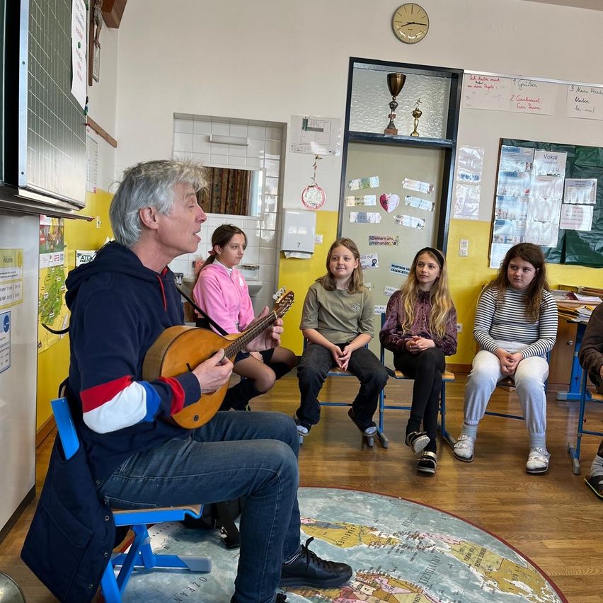 Ein älterer Mann spielt eine Laute, während vier Mädchen aufmerksam zuhören in einem Klassenzimmer. Der Raum ist mit Postern und Trophäen dekoriert.