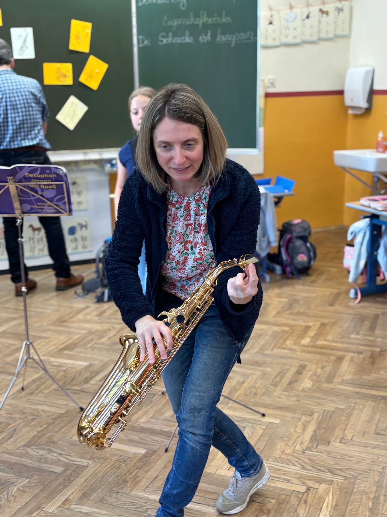 Eine Frau in einem Blumenhemd und Jeans hält eine Klarinette in einem Raum. Hinter ihr steht ein Mann in einem karierten Hemd und Lederschuhen in der Nähe einer Tafel.