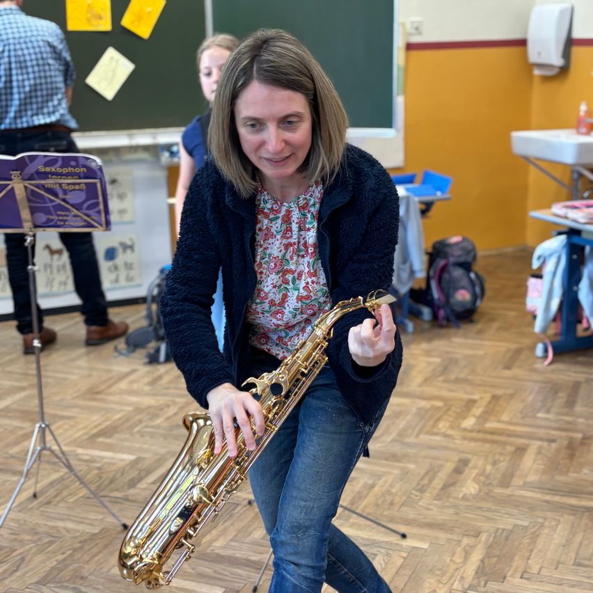 Eine Frau in einem Blumenhemd und Jeans hält eine Klarinette in einem Raum. Hinter ihr steht ein Mann in einem karierten Hemd und Lederschuhen in der Nähe einer Tafel.