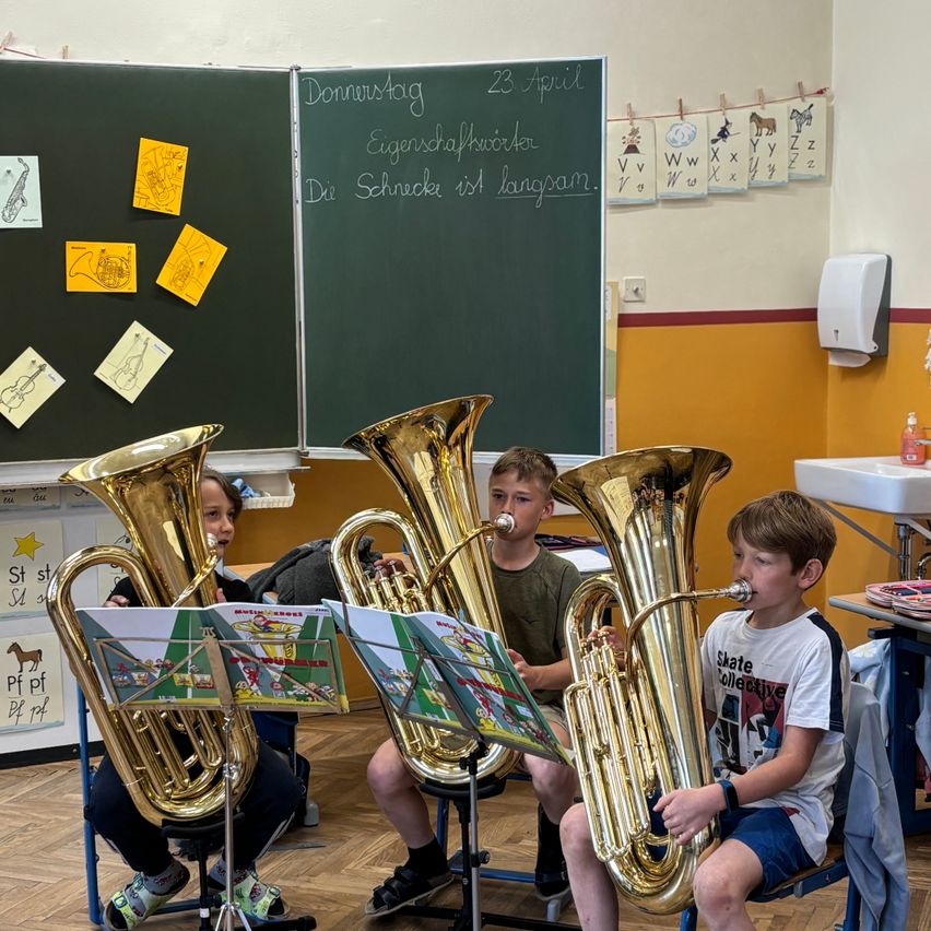 Vier Kinder spielen Tubas in einem Klassenzimmer. Die Tafel hinter ihnen hat Musikinstrumente und Noten angeheftet. Ein Waschbecken und Handdesinfektionsmittel befinden sich auf der rechten Seite.