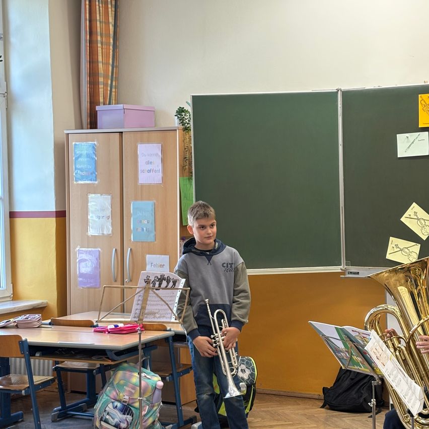 Ein junger Junge steht in einem Klassenzimmer und hält eine Trompete. Hinter ihm ist eine grüne Tafel an der Wand montiert, mit Klebenotizen. Ein Tisch mit einem Notenständer steht in der Nähe.