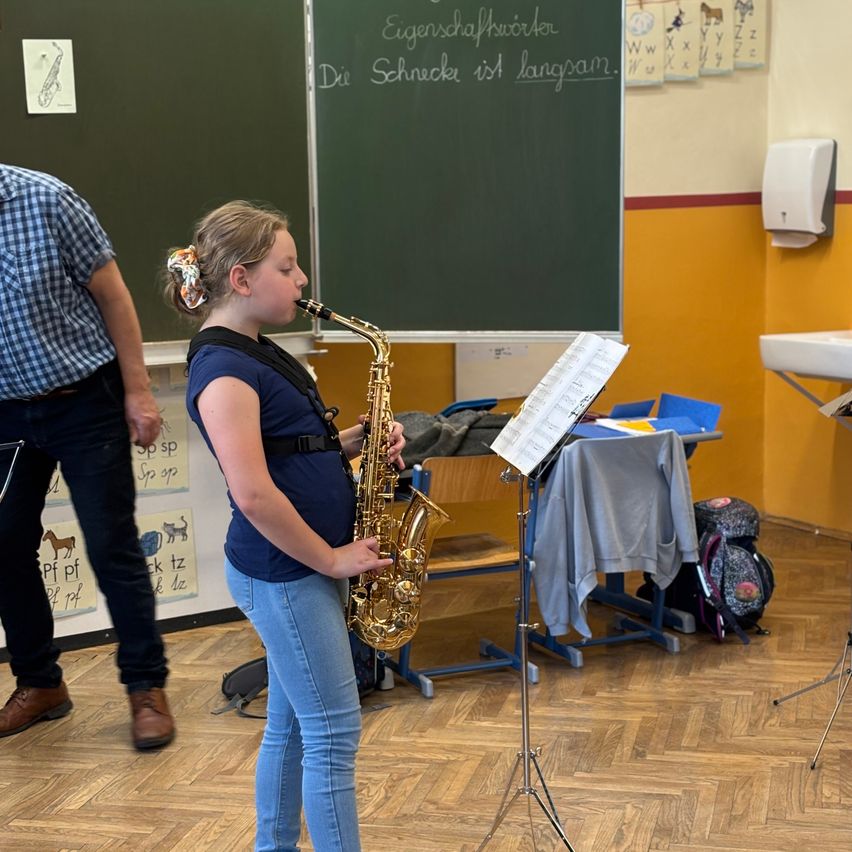 Ein junges Mädchen spielt Saxophon in einem Klassenzimmer, vor ihr steht ein Notenständer und ein Mann steht neben ihr. Hinter ihr zeigt eine Tafel deutsche Wörter.