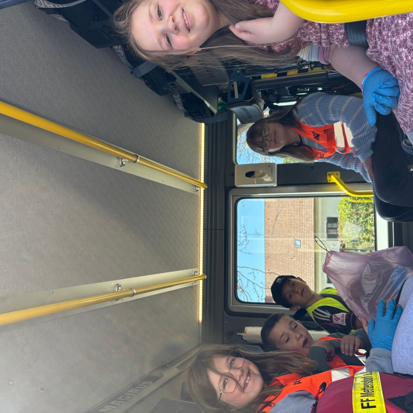 Mehrere Kinder und Erwachsene in Sicherheitswesten sitzen in einem Krankenwagen und lächeln. Ein Kind hält einen Plastikbeutel. Der Innenraum ist gelb mit Metall-Handläufen.