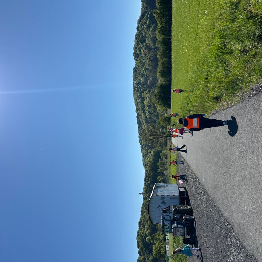 Eine Gruppe von Menschen in orangefarbenen Westen geht neben einem Lastwagen auf einem Pfad. Eine Person in Blau steht in der Nähe. Der Weg wird von einem Grasfeld und einem Berg flankiert.