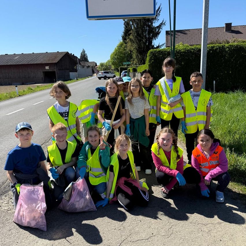 Eine Gruppe von Kindern in Warnwesten und Handschuhen posiert für ein Foto, während sie Mülltüten halten, vor einer Straße mit einem Schild und Häusern im Hintergrund.