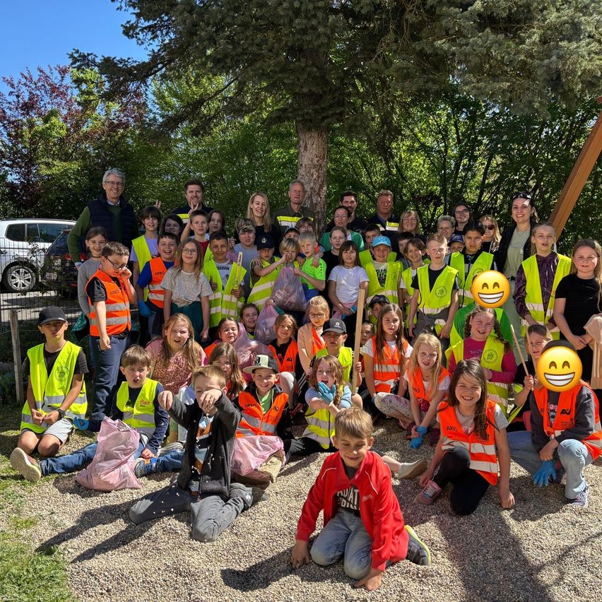 Eine Gruppe von Menschen, darunter Kinder, posiert für ein Foto im Park. Sie tragen hellgelbe Westen und einige halten Schilder. Hinter ihnen stehen Bäume und ein Auto.