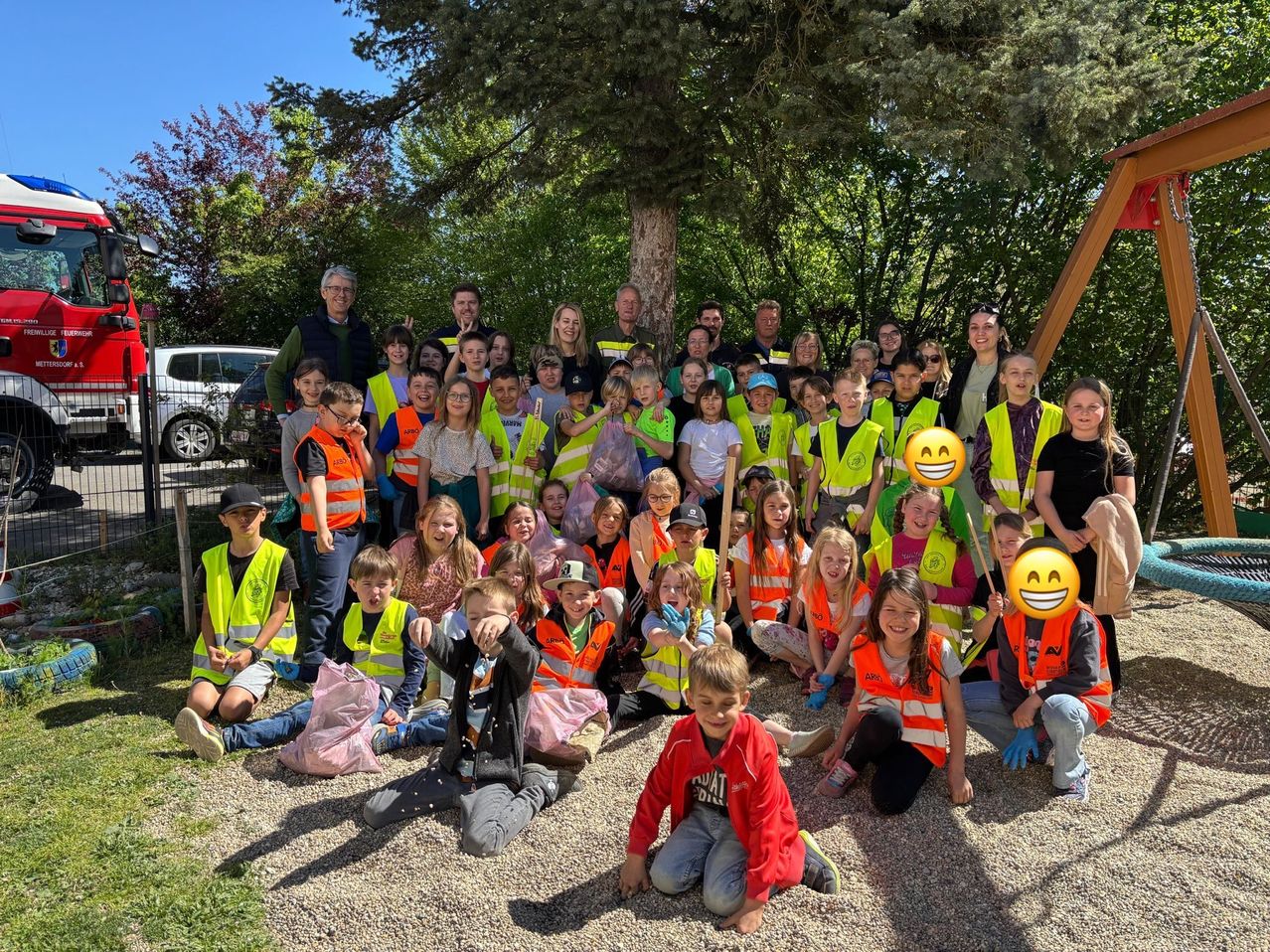 Eine Gruppe von Menschen, darunter Kinder, posiert für ein Foto im Park. Sie tragen hellgelbe Westen und einige halten Schilder. Hinter ihnen stehen Bäume und ein Auto.