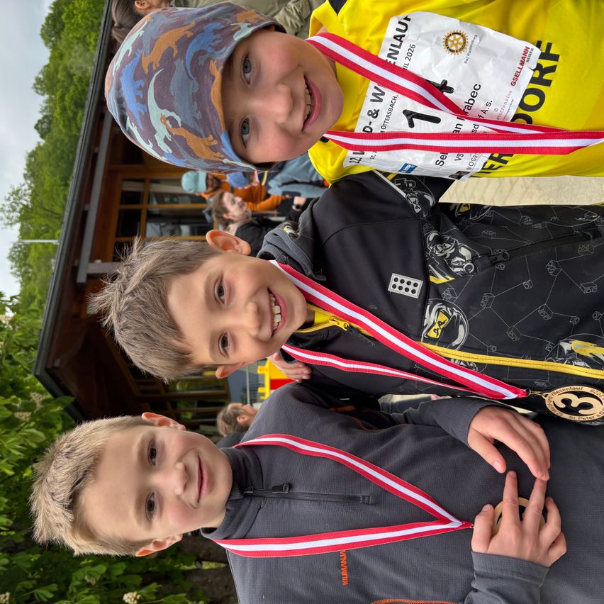 Drei Jungen mit Medaillen stehen eng beieinander und lächeln. Der Junge in der Mitte trägt ein gelbes Shirt mit einer Startnummer und Name. Der Junge auf der rechten Seite hat eine Camouflage-Mütze.