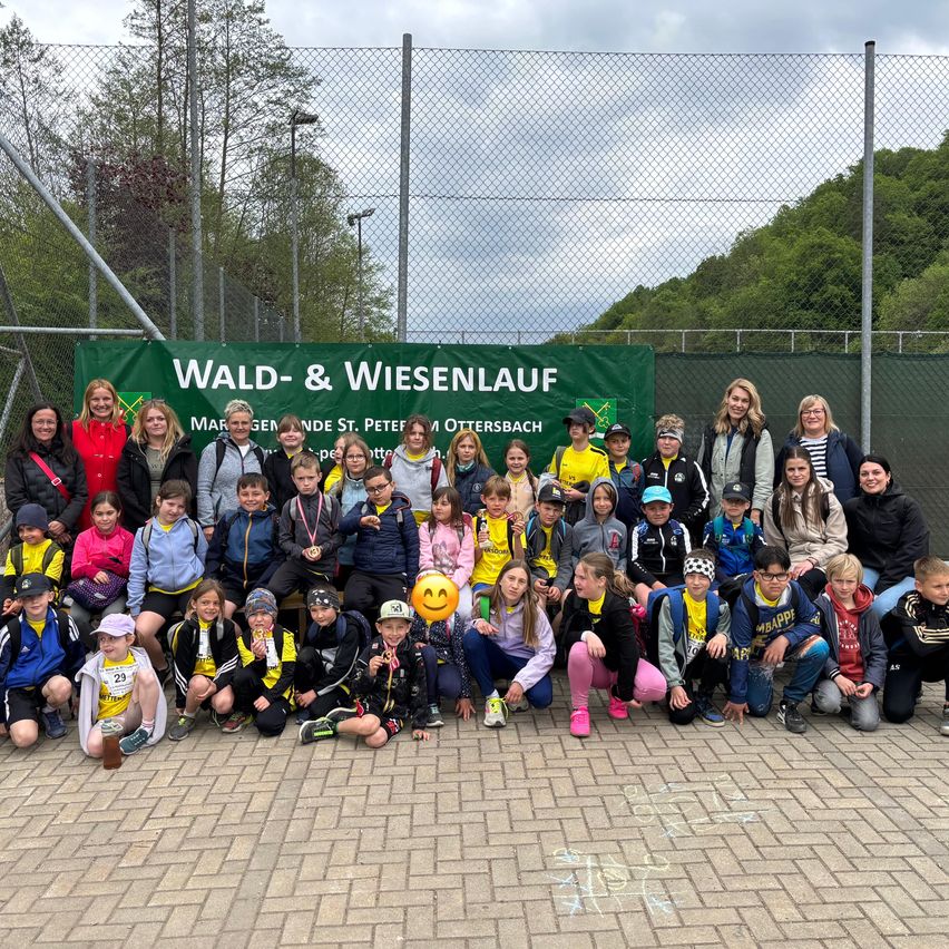Eine Gruppe von Kindern und Erwachsenen steht vor einem Zaun mit einem Banner, das Wald- & Wiesenlauf steht. Einige tragen Medaillen und alle tragen Freizeitkleidung. Dahinter befinden sich Bäume und ein Berg.