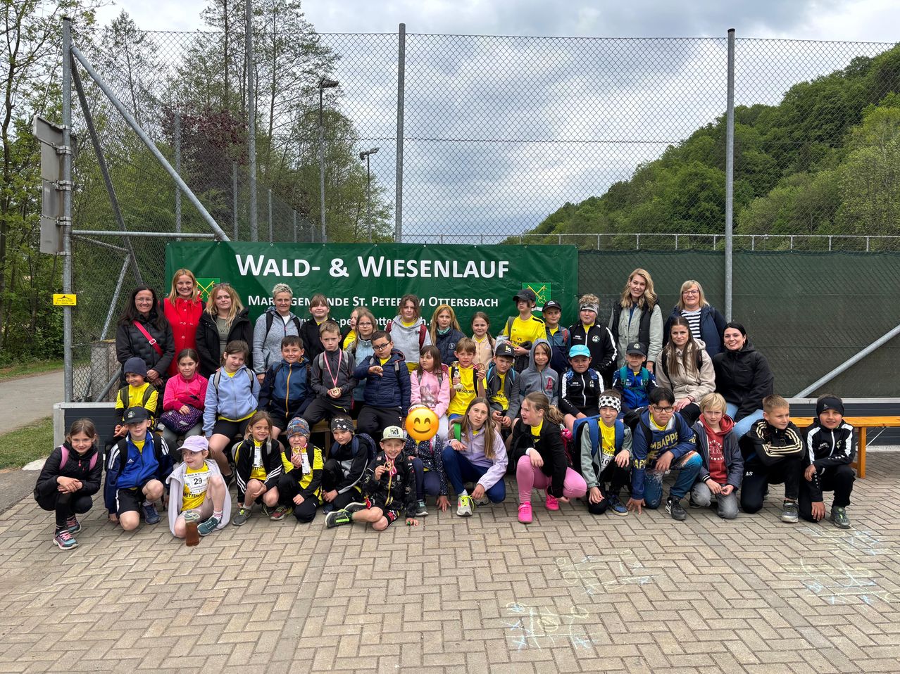 Eine Gruppe von Kindern und Erwachsenen steht vor einem Zaun mit einem Banner, das Wald- & Wiesenlauf steht. Einige tragen Medaillen und alle tragen Freizeitkleidung. Dahinter befinden sich Bäume und ein Berg.