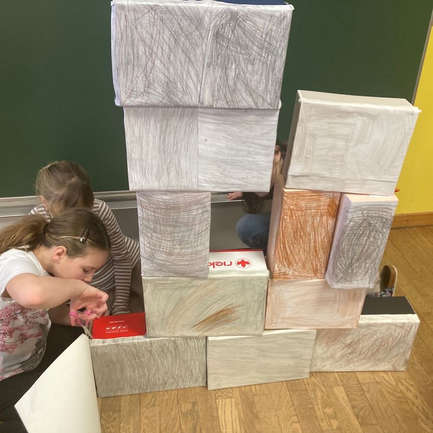 Zwei Mädchen bauen in einem Klassenzimmer eine Turm aus bunten Kisten. Sie tragen Freizeitkleidung.