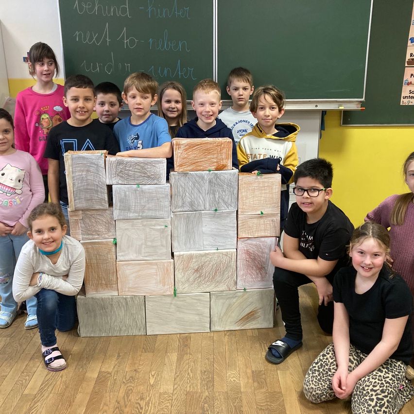 Eine Gruppe von Kindern posiert für ein Foto in einem Klassenzimmer. Sie stehen hinter einem Turm aus Holzklötzen. Dahinter hat eine Tafel etwas Schriftliches darauf.