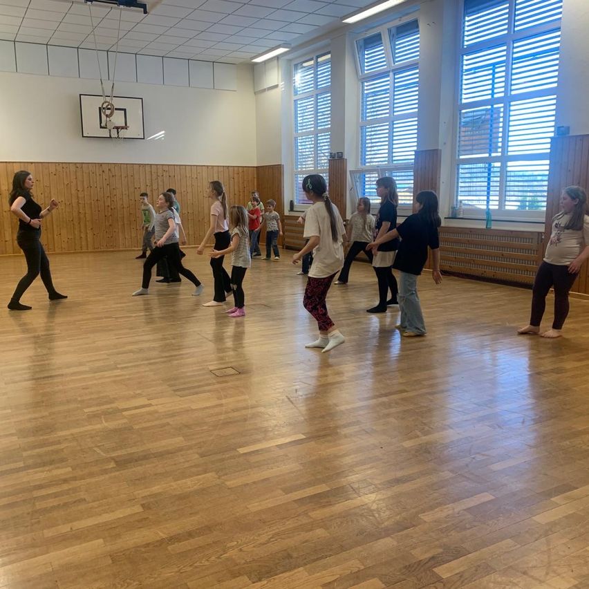 Eine Gruppe von Mädchen und Frauen übt Tanzschritte in einer Turnhalle mit Holzböden und großen Fenstern. Eine Frau leitet die Sitzung.