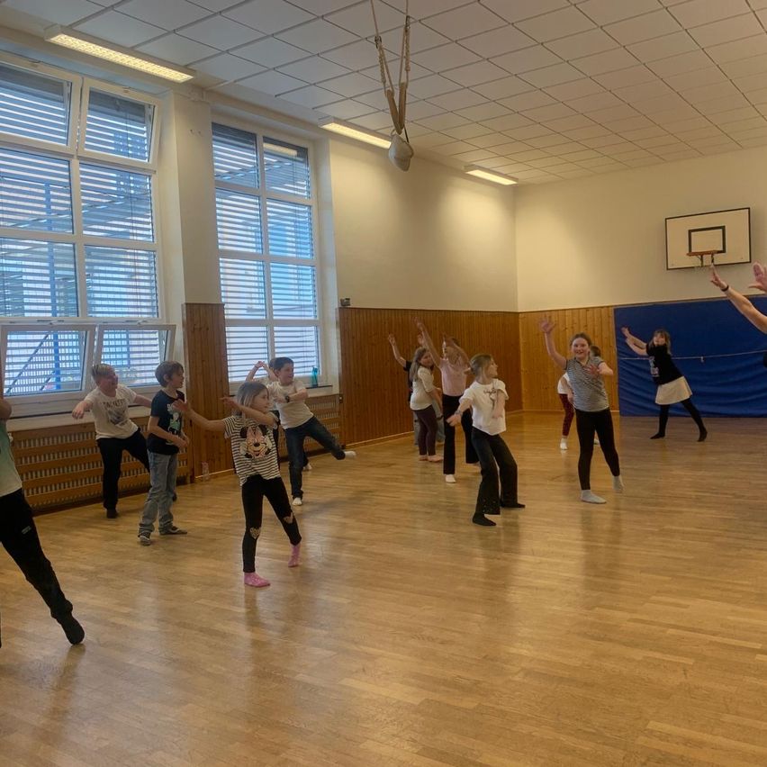 Eine Gruppe von Kindern tanzt in einer Turnhalle. Einige tragen Turnschuhe, andere sind barfuß. Sie machen verschiedene Posen.