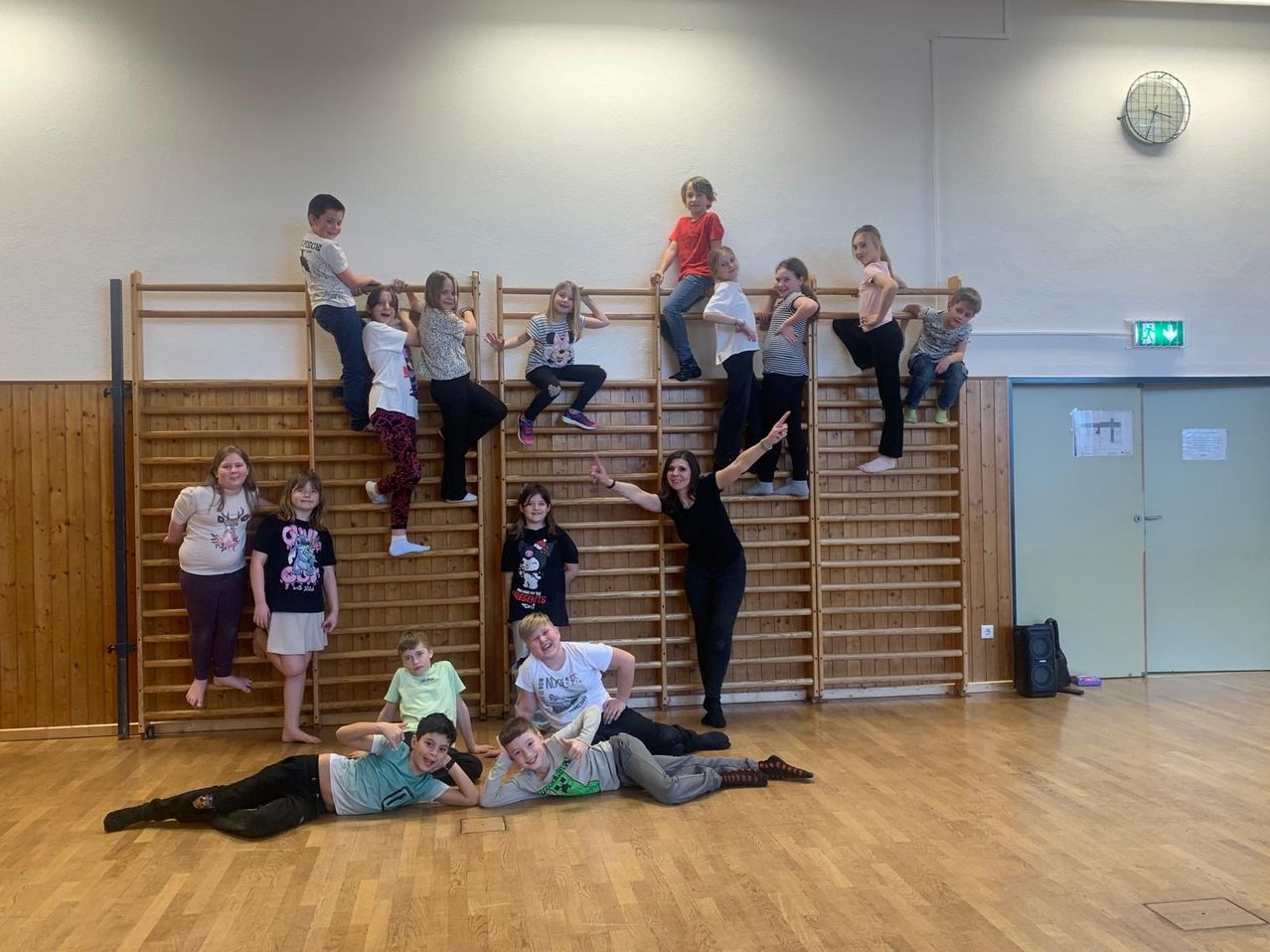 Eine Gruppe von Kindern und einer Frau befinden sich in einem Raum, einige klettern auf Holzstangen und andere sitzen auf dem Boden. Die Frau zeigt auf die Kinder an den Stangen.