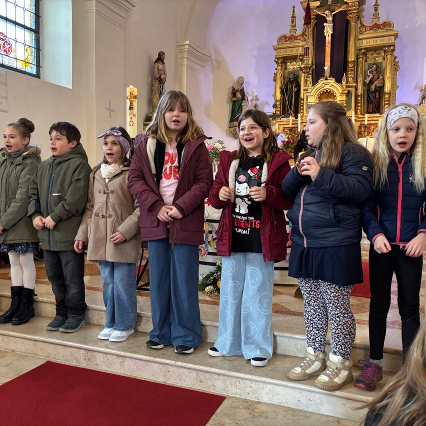 Eine Gruppe von Kindern steht auf den Stufen einer Kirche und lächelt. Die Kinder tragen Winterkleidung. Hinter ihnen stehen eine Statue und ein Kreuz auf dem Altar.