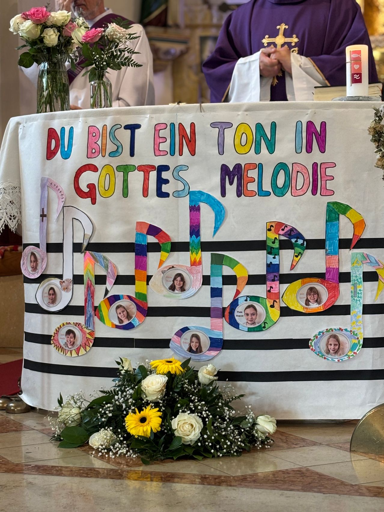 Ein Tisch ist mit einem Banner dekoriert, das in leuchtenden Farben 'Du bist ein Ton in Gottes Melodie' steht. Das Banner ist mit Musiknoten und Bildern von Menschen geschmückt. Ein weißer Blumenstrauß liegt am Fuß des Tisches.
