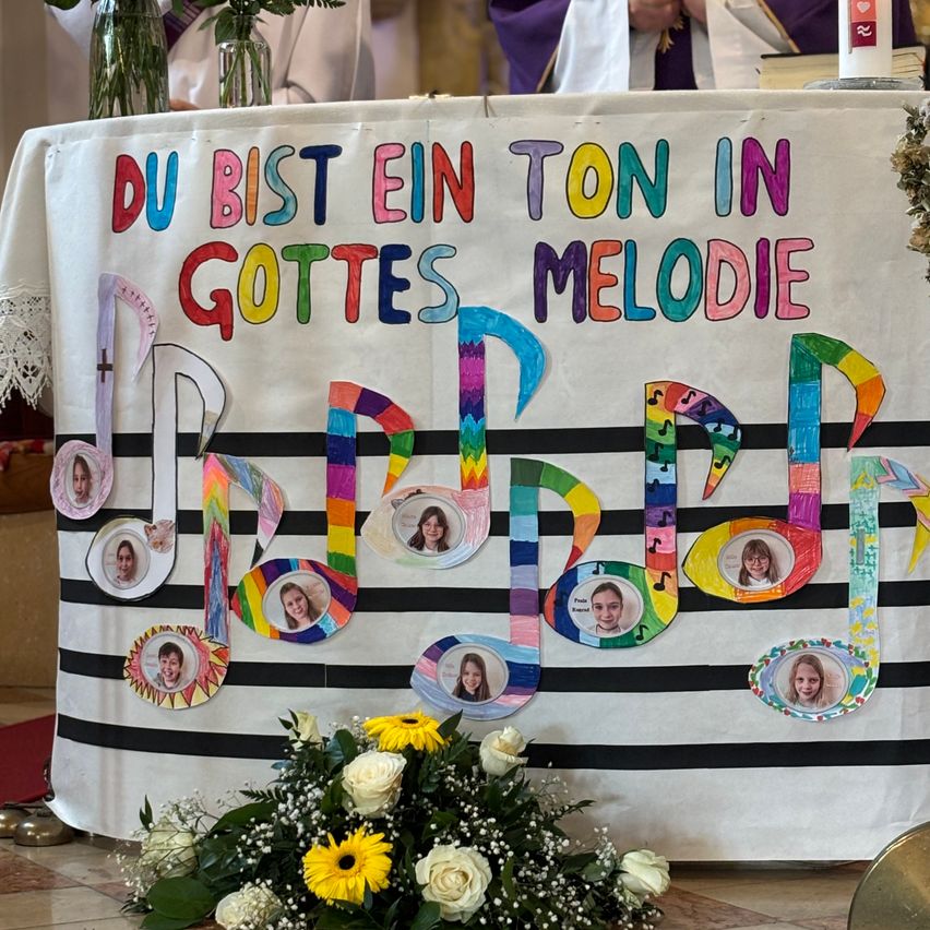 Ein Tisch ist mit einem Banner dekoriert, das in leuchtenden Farben 'Du bist ein Ton in Gottes Melodie' steht. Das Banner ist mit Musiknoten und Bildern von Menschen geschmückt. Ein weißer Blumenstrauß liegt am Fuß des Tisches.