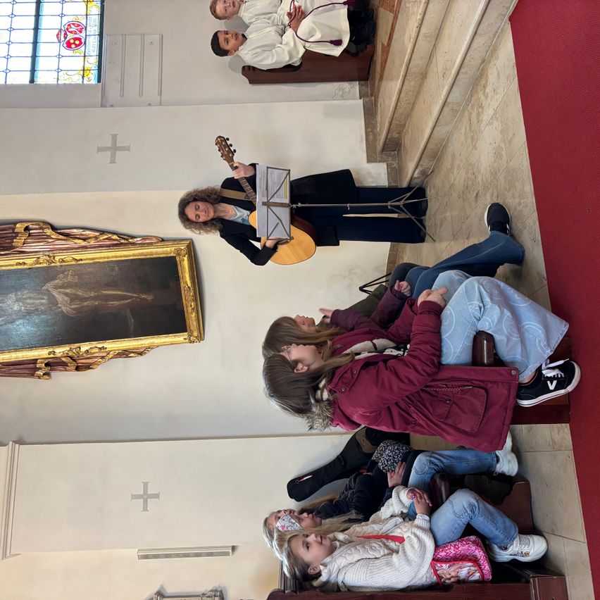 Eine Frau spielt Gitarre in einer Kirche, umgeben von Kindern auf den Stufen. Ein großes Gemälde schmückt die Wand oberhalb.