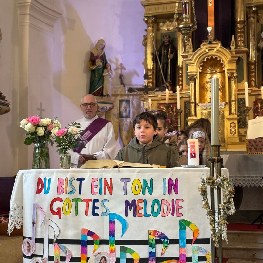 Ein junger Junge steht an einem Altar mit einem Banner, das 'Du bist ein Ton in Gottes Melodie' besagt. Er liest aus einem offenen Buch, während ein Priester hinter ihm zuhört. Kerzen und Blumen schmücken den Bereich.