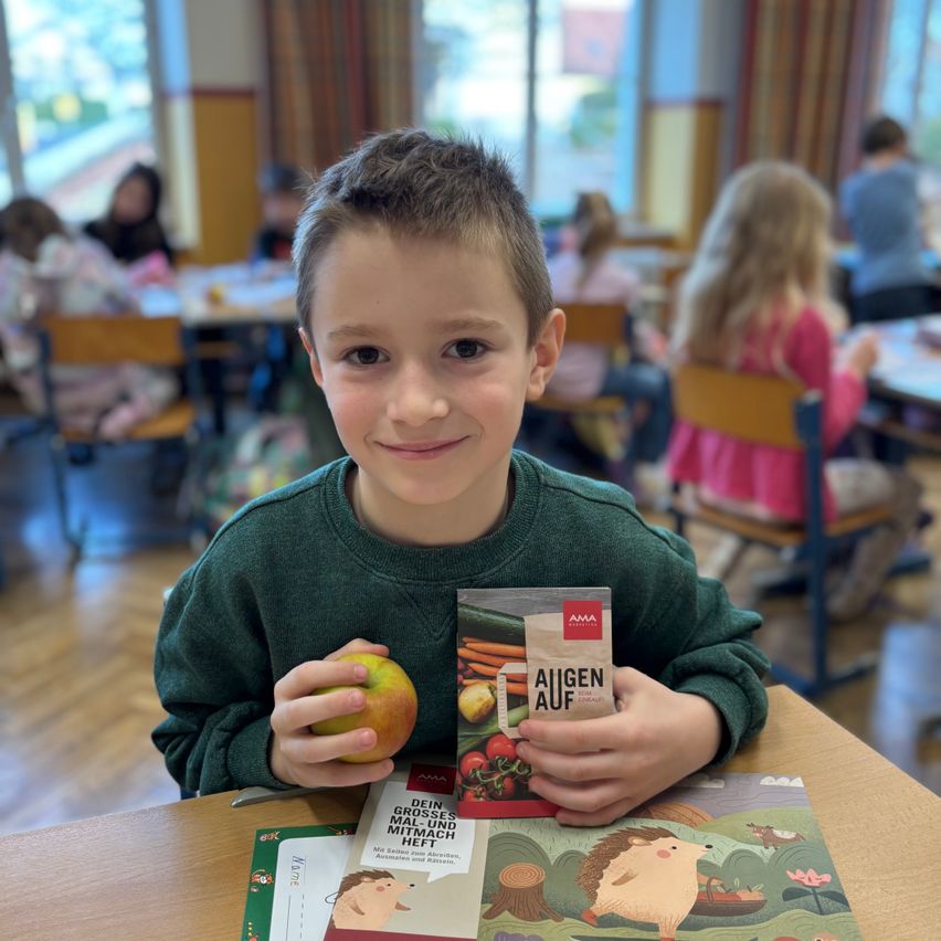 Ein Junge in einem grünen Sweatshirt hält einen Apfel und eine Tüte Samen in einem Klassenzimmer. Andere Kinder sitzen im Hintergrund an Tischen.