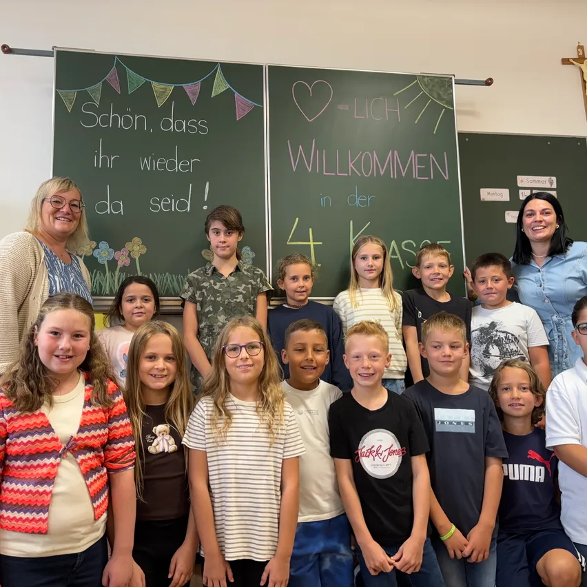 Eine Gruppe von Kindern und zwei Frauen stehen vor einem Klassenzimmer mit zwei Tafeln. Eine Tafel sagt 'Schon, dass ihr wieder da seid!' und die andere sagt 'Willkommen in der 4. Klasse.' Die Kinder lächeln.