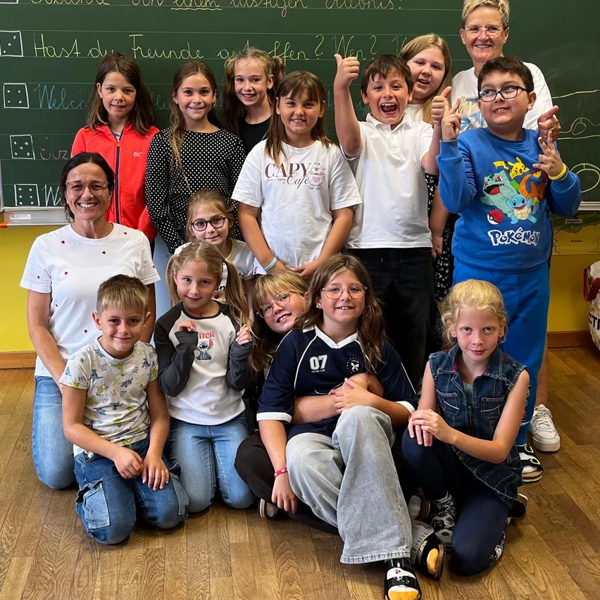 Eine Gruppe von Kindern und zwei Erwachsenen posiert für ein Foto im Klassenzimmer. Die Kinder tragen Freizeitkleidung und einige lächeln. Die Tafel hinter ihnen hat einige Text darauf geschrieben.