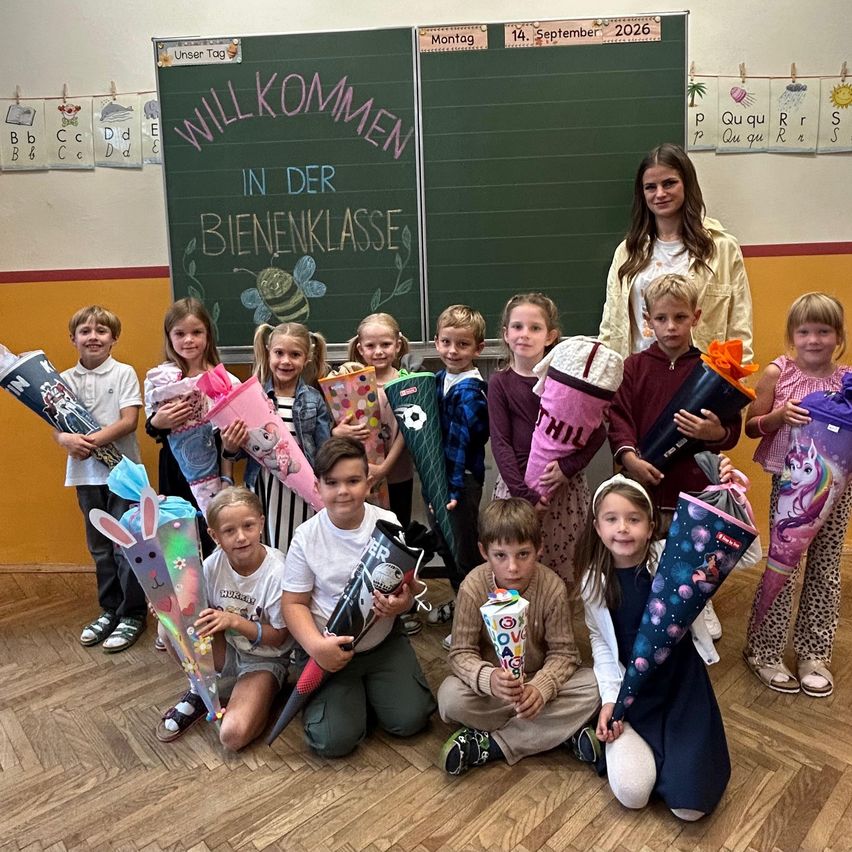 Eine Gruppe von Kindern und einer Frau posiert für ein Foto im Klassenzimmer. Die Kinder halten Blumensträuße und die Frau steht hinter ihnen. Die Tafel hat eine Willkommensbotschaft und ein Datum.