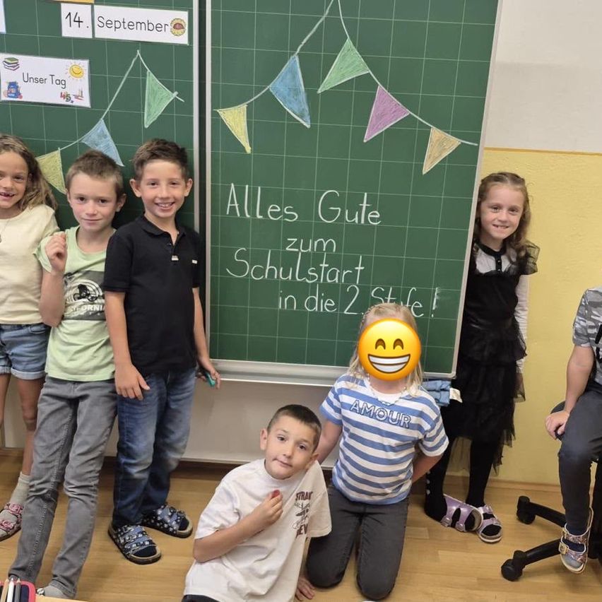 Eine Gruppe von Kindern posiert für ein Foto vor einer Tafel mit einer fröhlichen Botschaft. Die Tafel steht 'Alles Gute zum Schulstart in die 2. Stufe!' mit bunten Wimpelgirlanden.