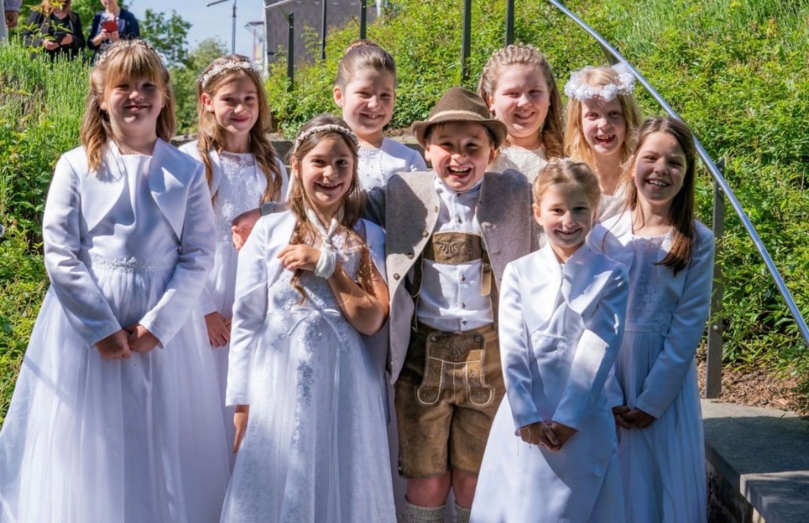 Eine Gruppe von Kindern in weißen Kleidern und ein Junge in traditioneller deutscher Kleidung stehen zusammen auf einer Treppe und lächeln für ein Foto.