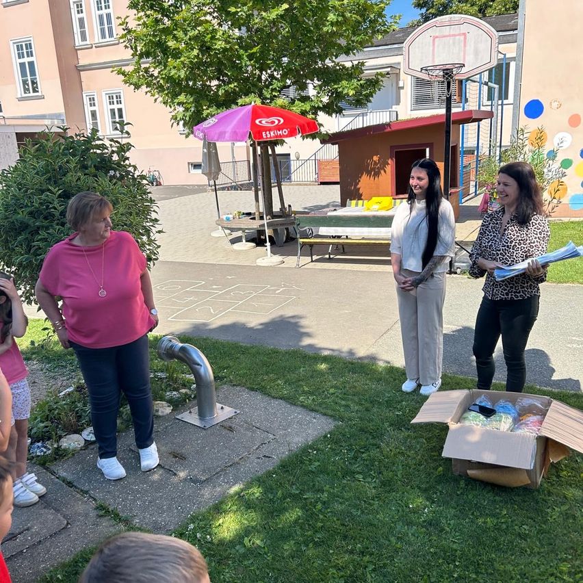 Drei Frauen stehen draußen in einem Park, zwei mit Papieren, eine hält eine Box. Dahinter steht ein Gebäude mit einem Basketballkorb und einem Baum.
