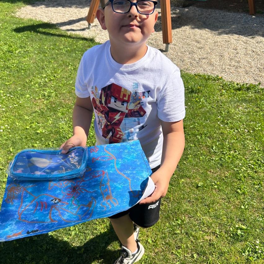 Ein Junge in einem weißen T-Shirt hält eine blaue Tasche mit Katzendesign auf einem Grasfeld. Er trägt eine Brille und schwarze Shorts.