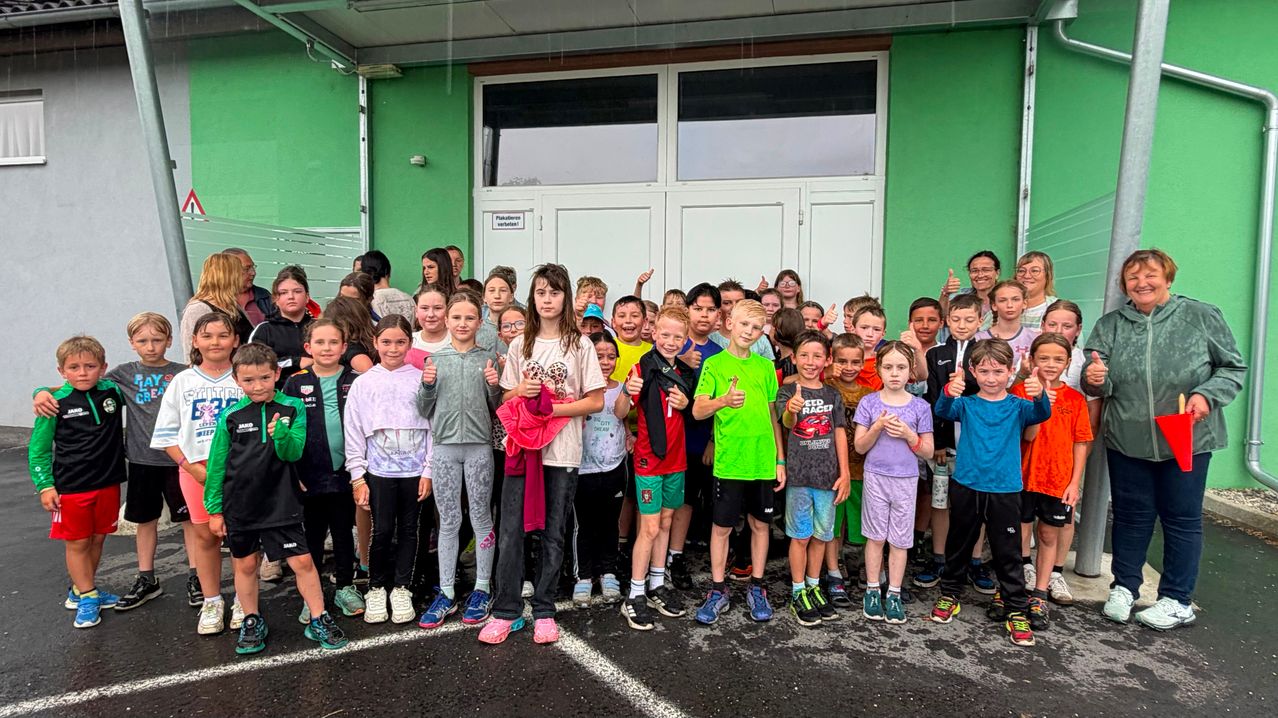 Eine Gruppe von Kindern und Erwachsenen posiert für ein Foto vor einem grünen Gebäude. Sie tragen verschiedene farbige Kleidung und Sneakers, lächeln und geben Daumen hoch.