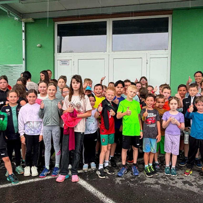 Eine Gruppe von Kindern und Erwachsenen posiert für ein Foto vor einem grünen Gebäude. Sie tragen verschiedene farbige Kleidung und Sneakers, lächeln und geben Daumen hoch.