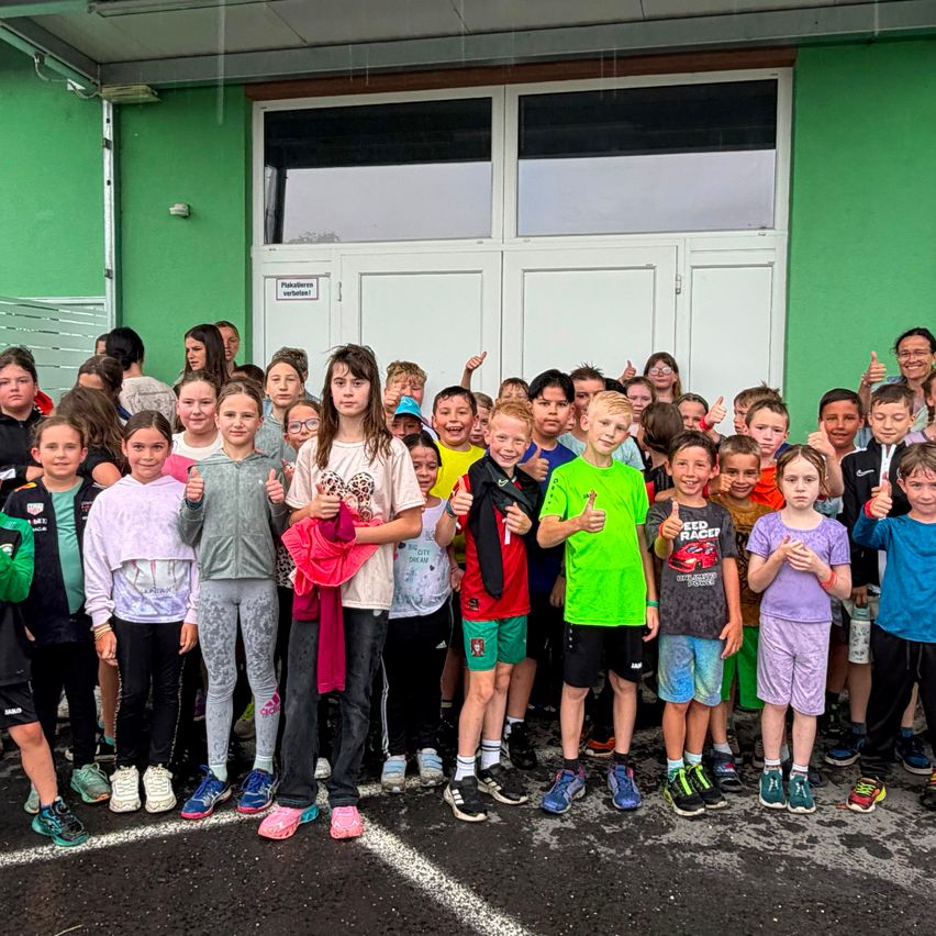 Eine Gruppe von Kindern und Erwachsenen posiert für ein Foto vor einem grünen Gebäude. Sie tragen verschiedene farbige Kleidung und Sneakers, lächeln und geben Daumen hoch.