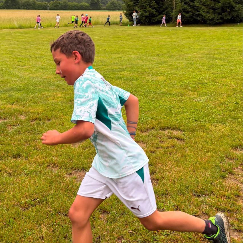 Ein junger Junge in einem Tie-Dye-Shirt und weißen Shorts läuft über ein Grasfeld. Dahinter laufen weitere Kinder in einem Rennen, während andere im Hintergrund stehen.