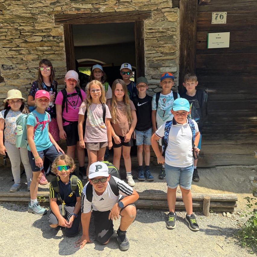 Eine Gruppe von Kindern in Hüten und Brillen posiert für ein Foto vor einem Steingebäude mit einem Schild, das 'Schule' steht.