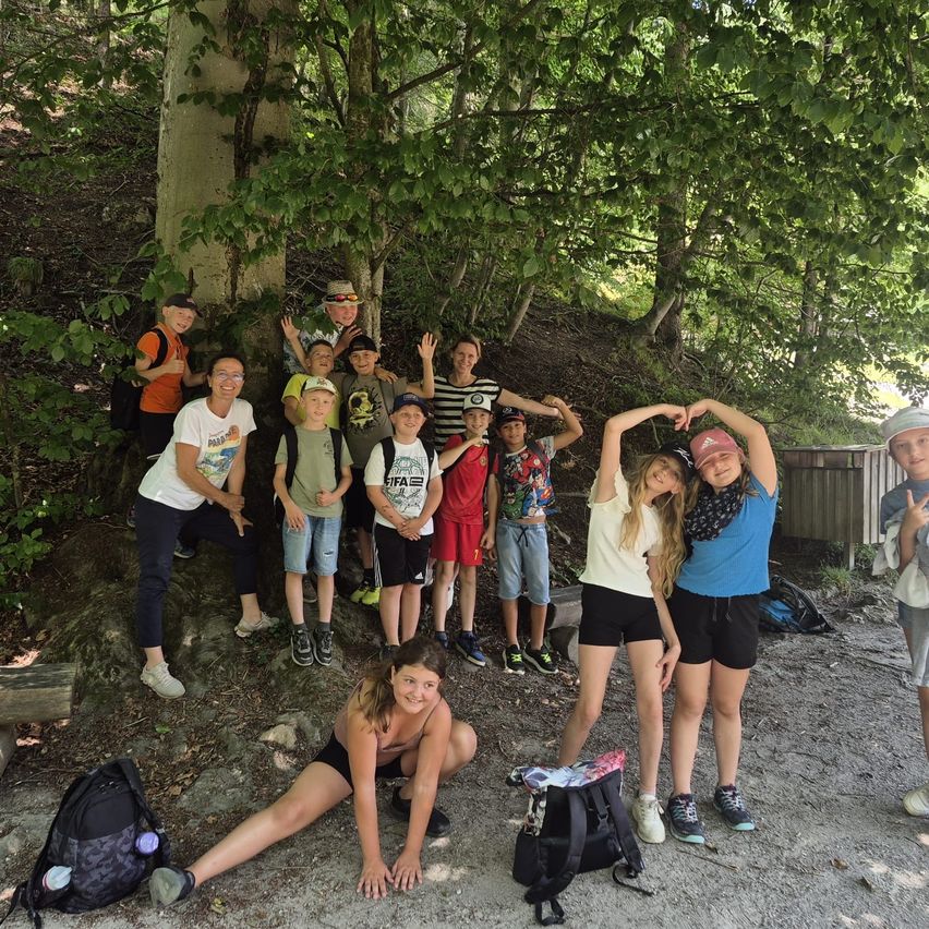 Eine Gruppe von Kindern und Erwachsenen posiert für ein Foto in einem bewaldeten Bereich, einige sitzen auf einem Felsen, andere stehen, mit Rucksäcken und einer Holzbank in der Nähe.